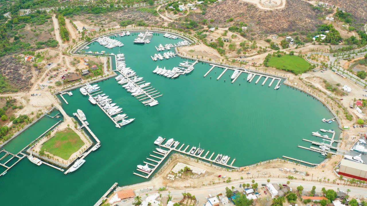 Aerial view across La Playita Los Cabos marina with docked yachts, empty slips, turquoise-green water, dry land, scattered buildings, and no visible people.