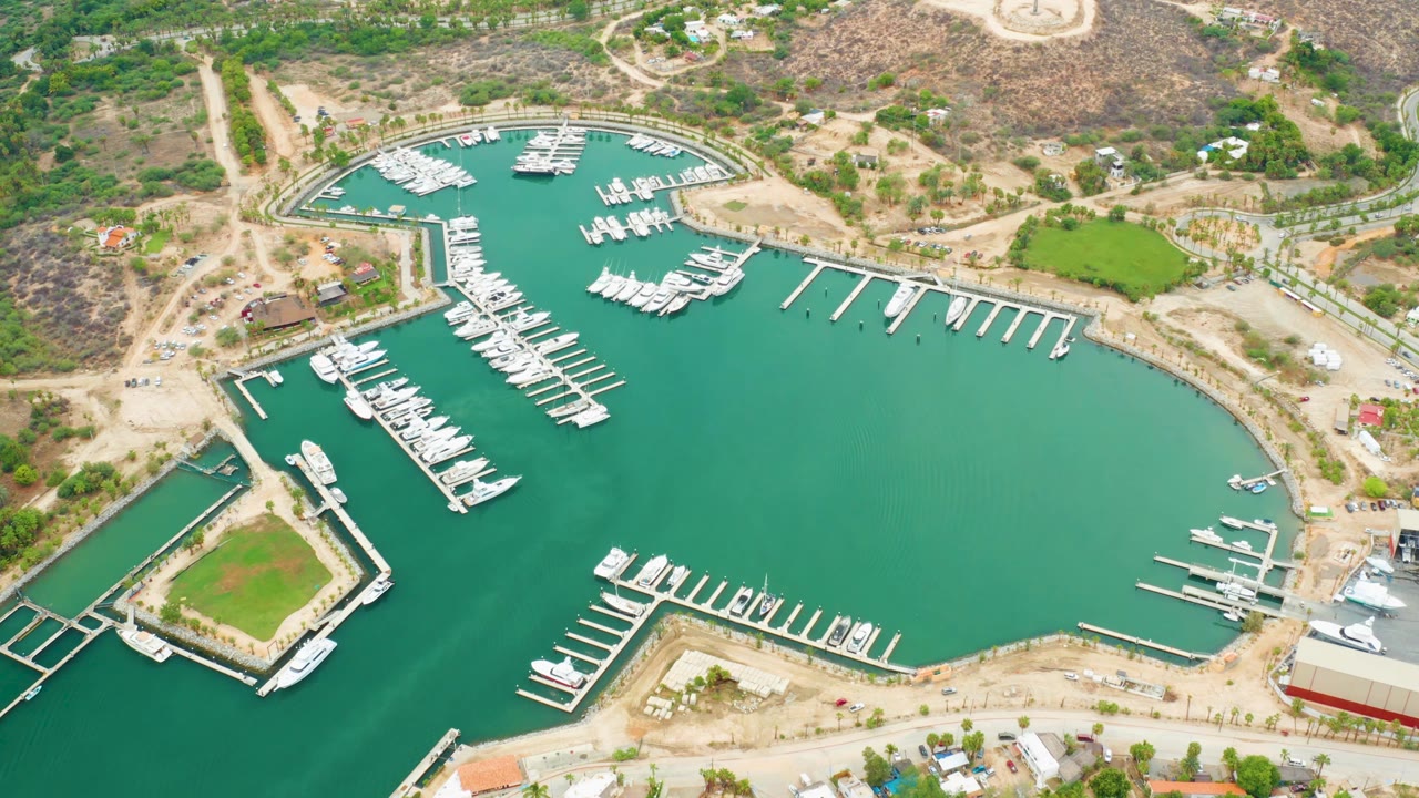 Aerial frame over La Playita Los Cabos marina showing green water, white boats along docks, dry hills, roads, scattered buildings, and no visible people.