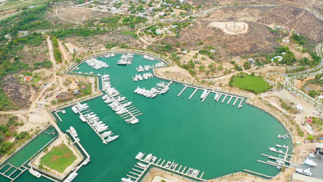 High drone-style view of La Playita Los Cabos marina with boats lined along docks, turquoise-green water, shore roads, dry hills, scattered buildings, and no visible people.