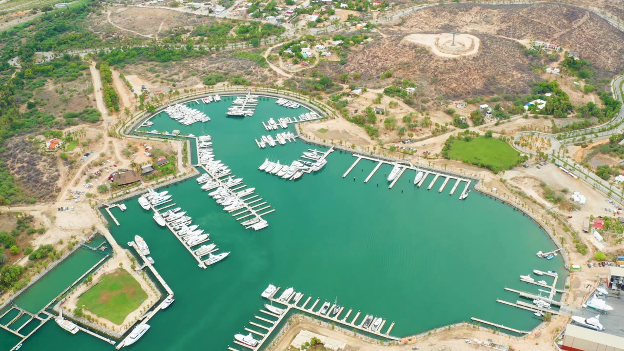 Aerial view in La Playita Los Cabos with the curved marina basin filling the frame, white boats at dock fingers, shore paths, dry hills, and no visible people.