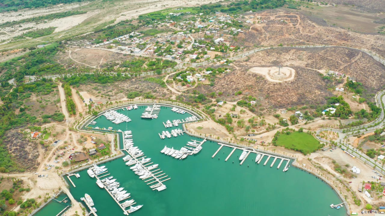 Opening aerial frame over La Playita Los Cabos showing turquoise marina water, white docks, clustered boats, curved roads, hillside homes, dry terrain, and no visible people.