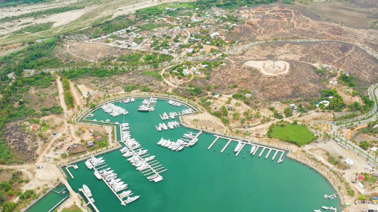 Aerial frame over La Playita Los Cabos showing more open marina water at the bottom, rows of slips and boats near center, dusty roads, scattered homes, dry hills, and no visible people.