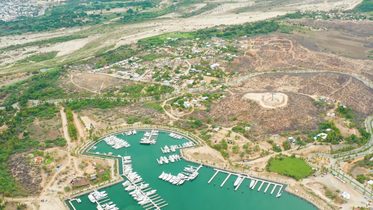 Aerial view over La Playita Los Cabos with white boats docked in a turquoise marina below dry hills, dirt roads, scattered buildings, and no visible people.