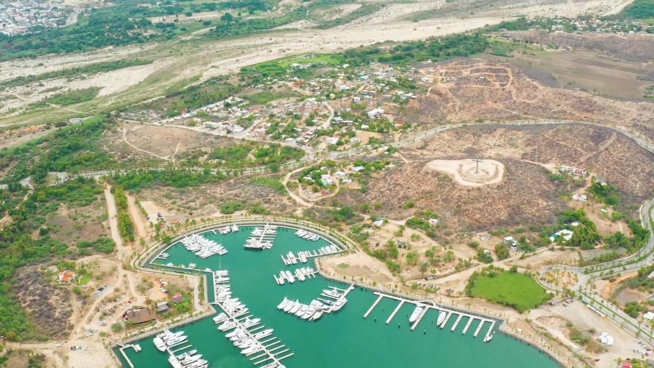 Drone-style aerial of La Playita Los Cabos with the marina across the lower frame, white boats lined along docks, tan hills and roads above, and no visible people.