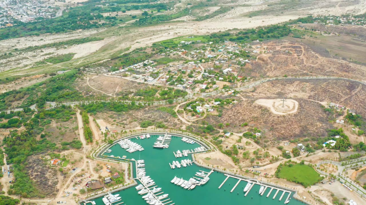 Aerial frame over La Playita Los Cabos showing turquoise marina water, white docked boats, dry hills, dirt roads, scattered buildings, and no visible people.