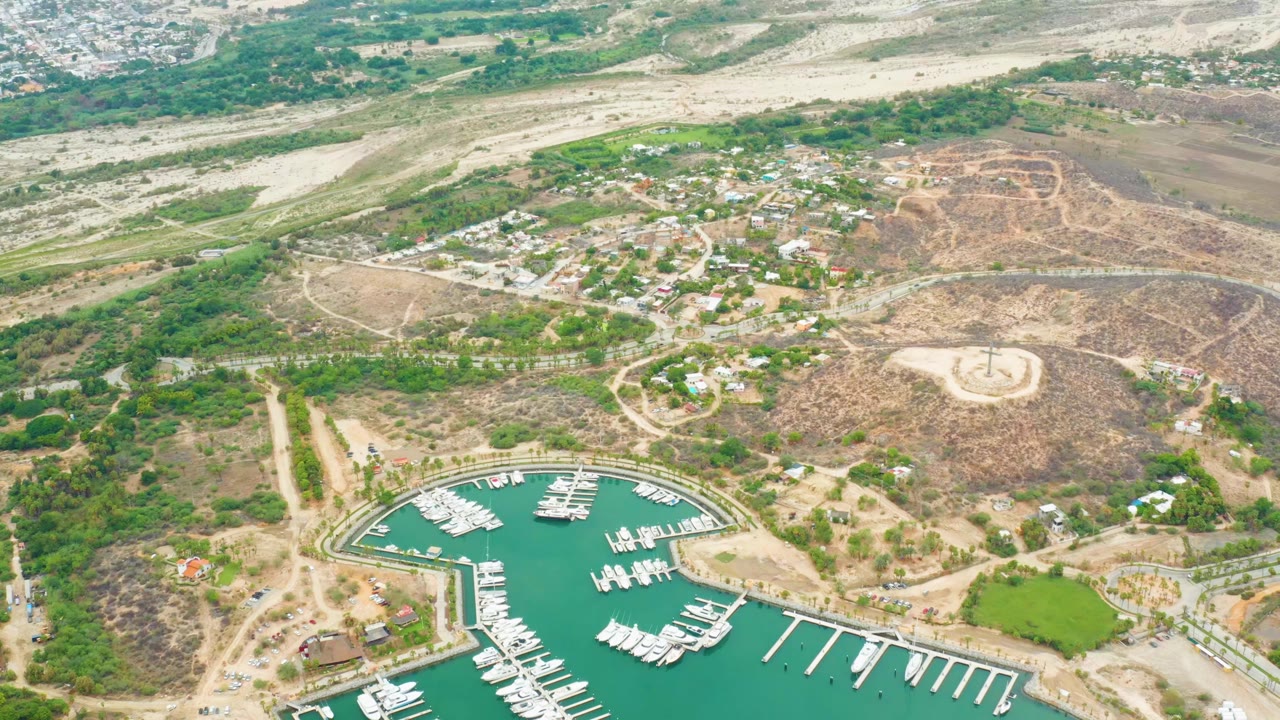 Aerial view in La Playita Los Cabos with rows of white boats in the marina, long piers, brown hills, roads, homes, and no visible people.