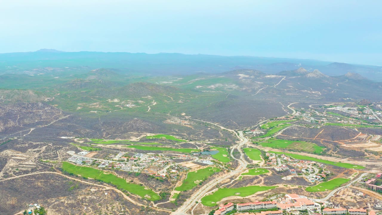 Opening aerial view over La Playita Los Cabos, with green fields, pale roads, scattered buildings, dry hills, hazy mountains, and no visible people.