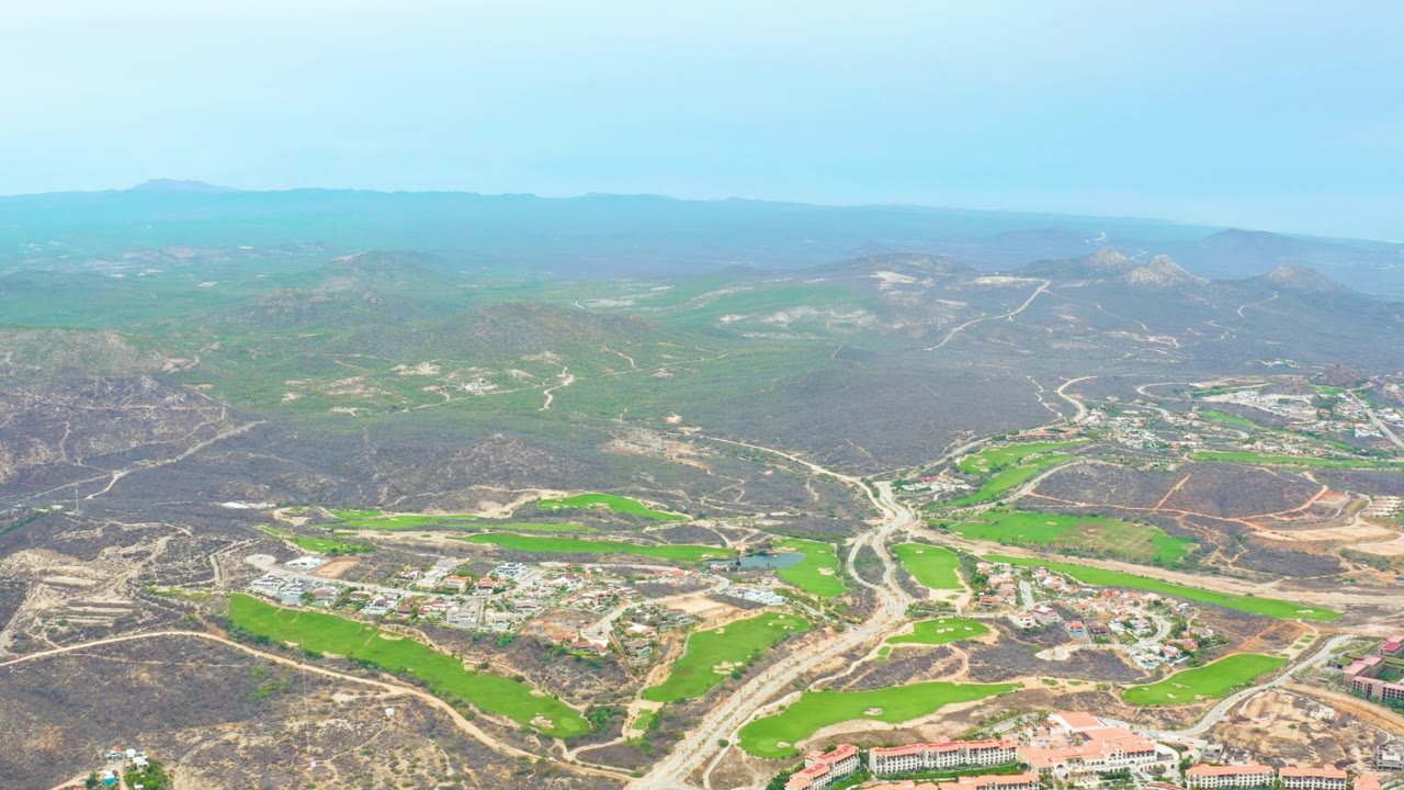 Early aerial frame over La Playita Los Cabos, showing the green valley floor, winding roads, clustered buildings, dry surrounding hills, and no visible people.
