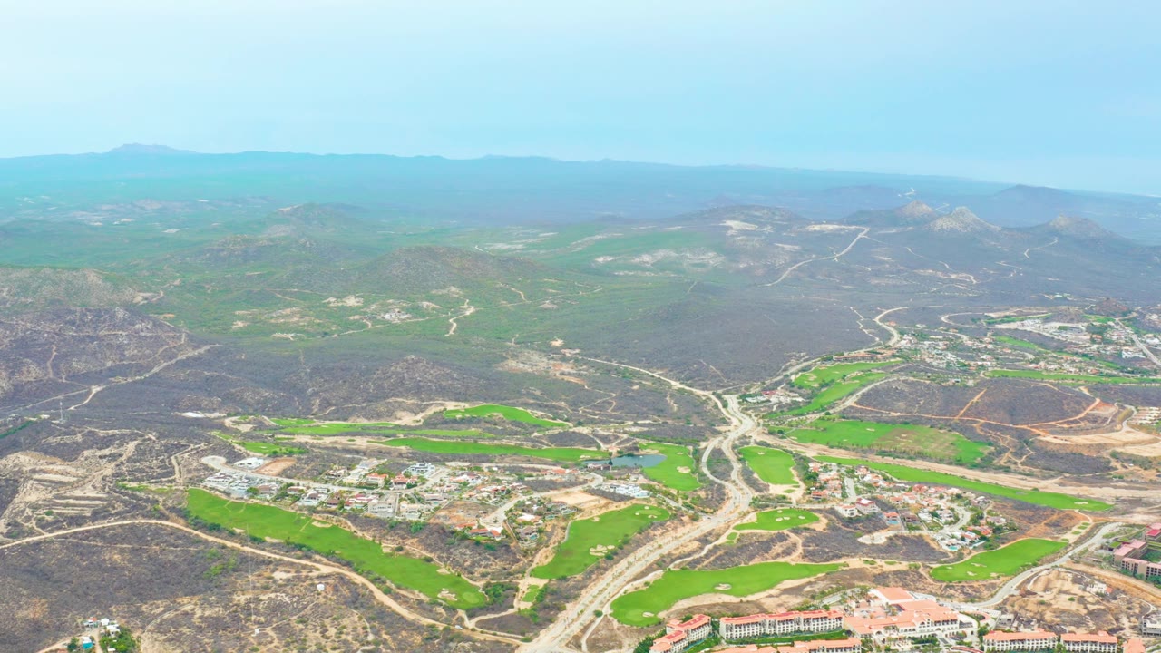 La Playita Los Cabos aerial frame with bright green fields in the lower valley, roads leading toward buildings, hazy mountain ridges, and no visible people.
