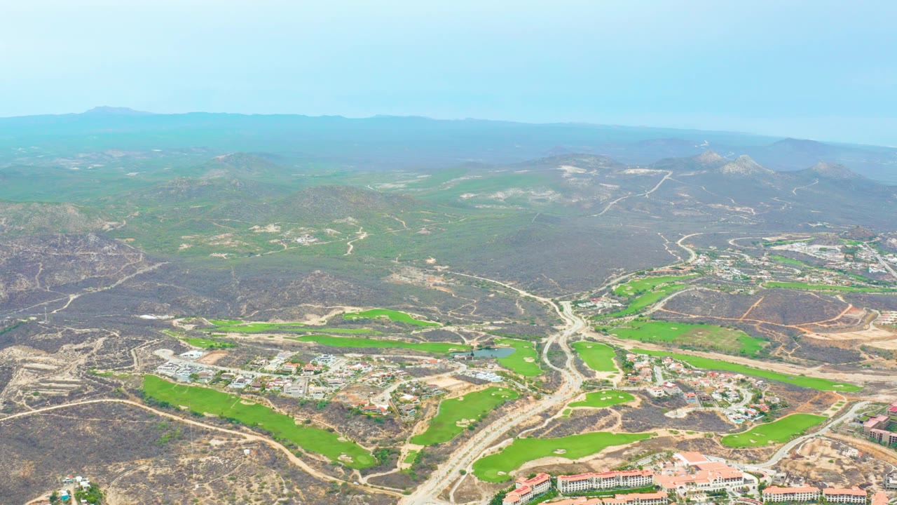 Aerial view across green fairway-like fields, road cuts, buildings, and brown hills.