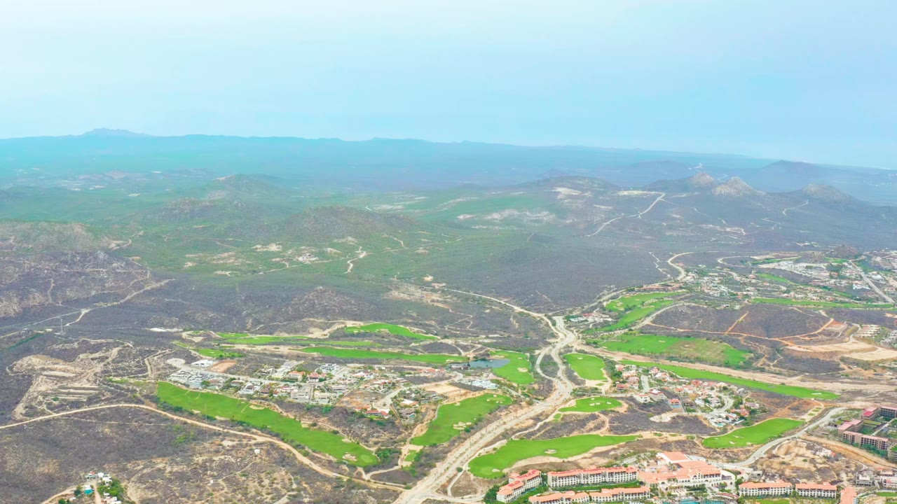 High aerial frame over La Playita Los Cabos with pale roads cutting through green fields, dry brown slopes, low buildings, distant ridgelines, and no visible people.