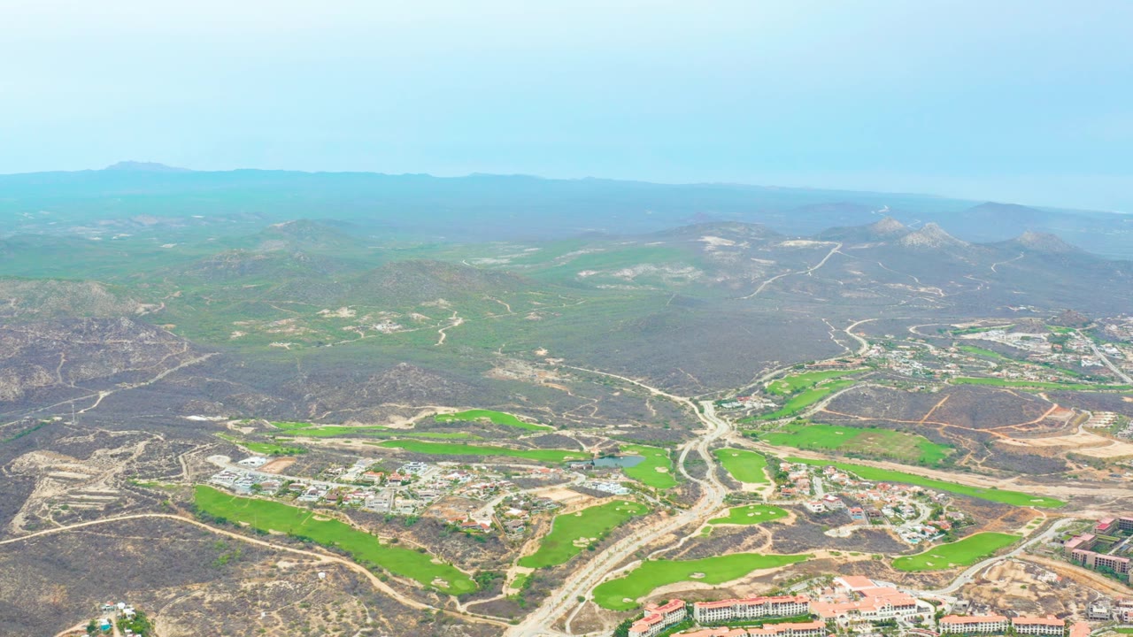Mid-clip aerial frame over La Playita Los Cabos with vivid green field areas, winding roads, brown hillside terrain, scattered structures, haze at the horizon, and no visible people.