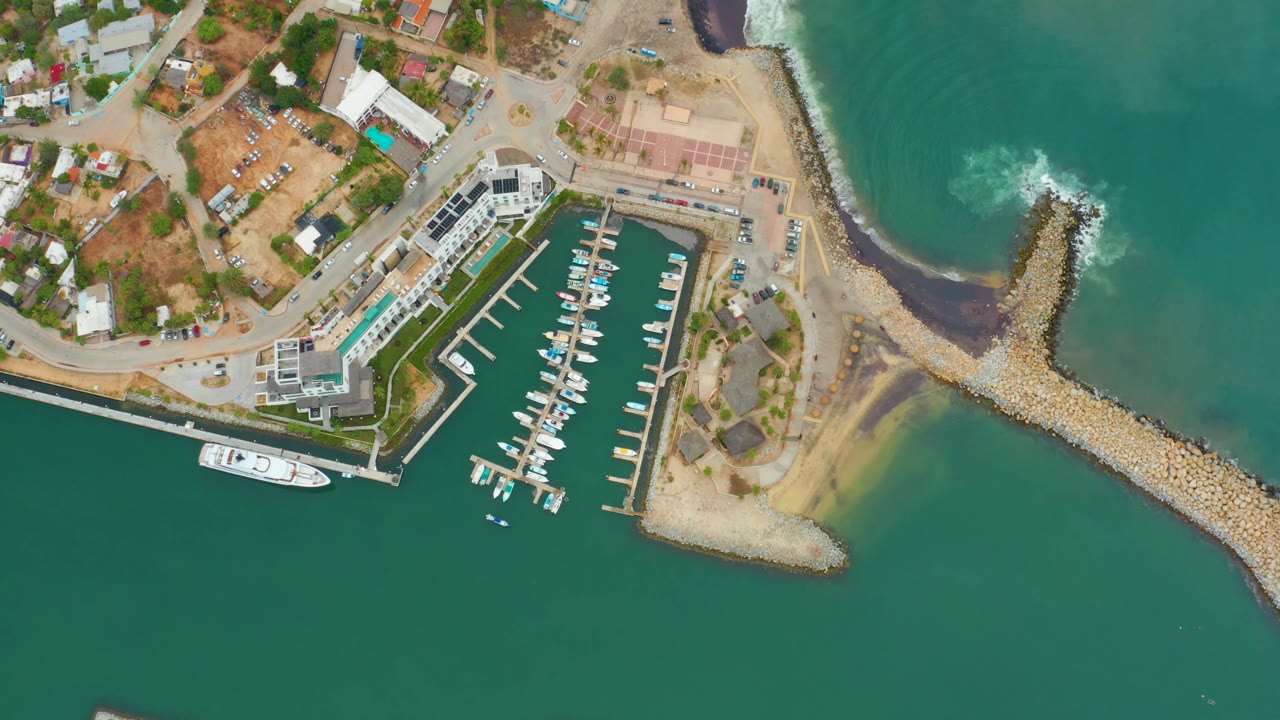 La Playita Los Cabos marina seen from above as the white boat sits farther from the dock edge, with foamy water striking the rocky breakwater and no visible people.