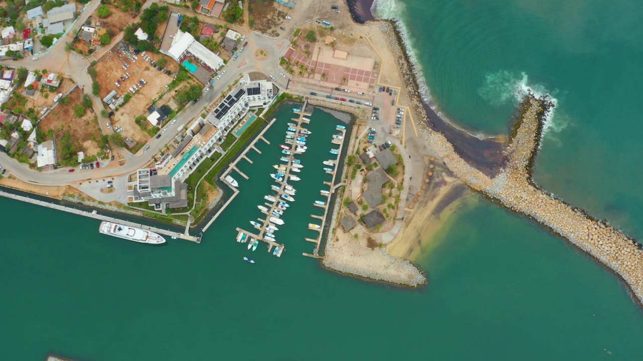 Top-down frame of the La Playita Los Cabos marina with rows of docked boats, the breakwater curving into green-blue water, fresh white surf foam, and no visible people.