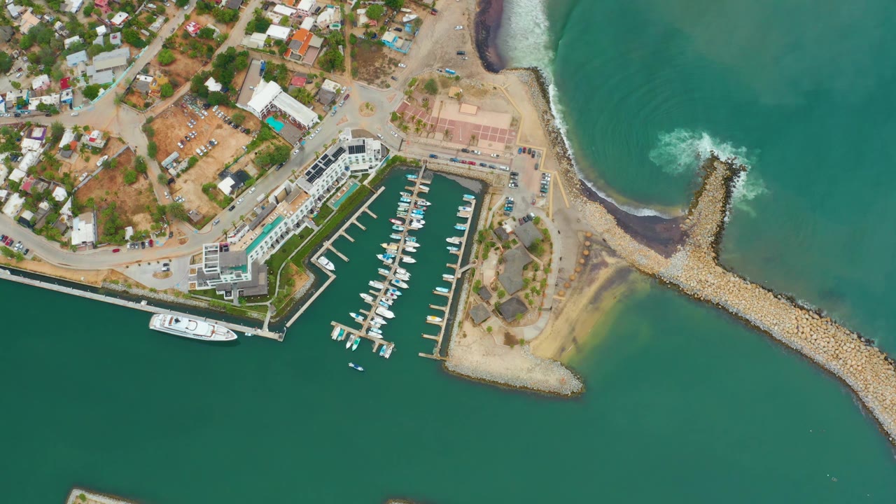 Aerial frame of La Playita Los Cabos showing dock slips, moored boats, coastal roads, dry shoreline, and white water forming at the breakwater with no visible people.