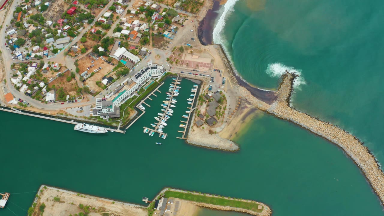 High aerial frame of La Playita Los Cabos with town structures above the marina, rows of small boats in slips, a white yacht at the quay, green harbor water, and no visible people.