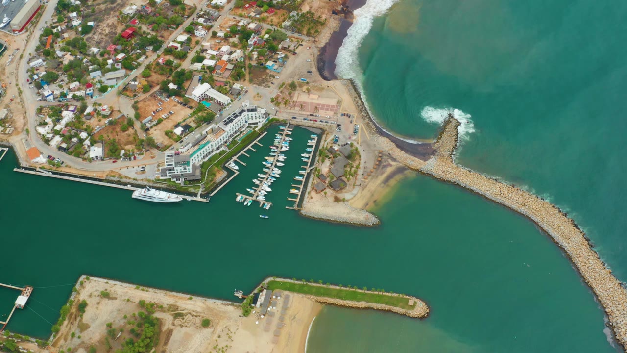 Opening overhead frame of La Playita Los Cabos with the marina docks near the upper center, green water around two rock breakwaters, a large white yacht, shore buildings, and no visible people.