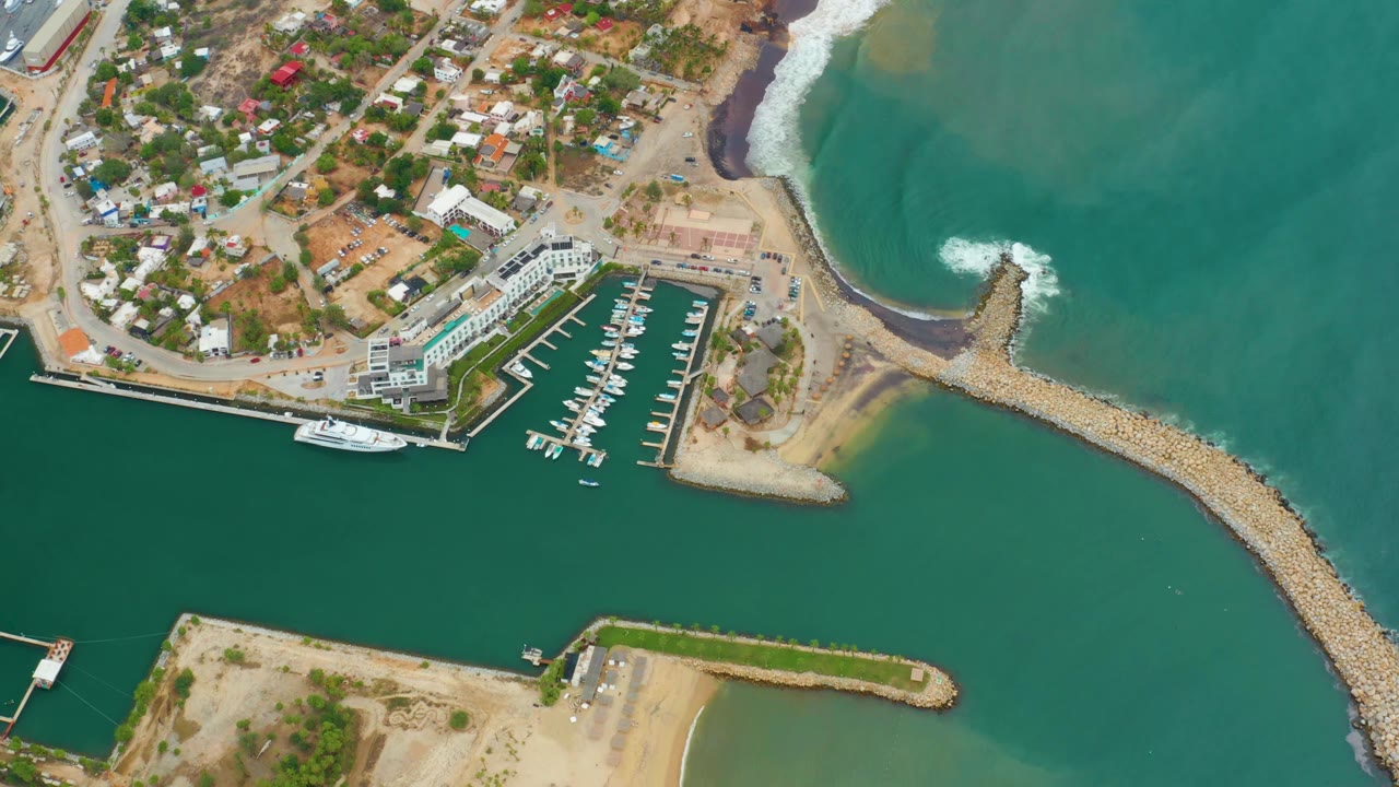 Second overhead frame of La Playita Los Cabos showing the white dock grid, docked boats, curved rock breakwater, sandy shore, nearby roads and buildings, and no visible people.