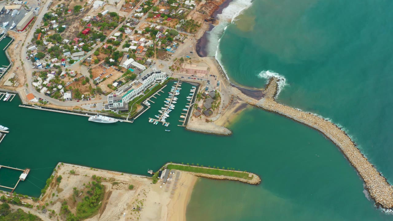 Aerial frame over La Playita Los Cabos with the marina centered, the breakwater curving through the right side, teal ocean beyond it, coastal homes above, and no visible people.