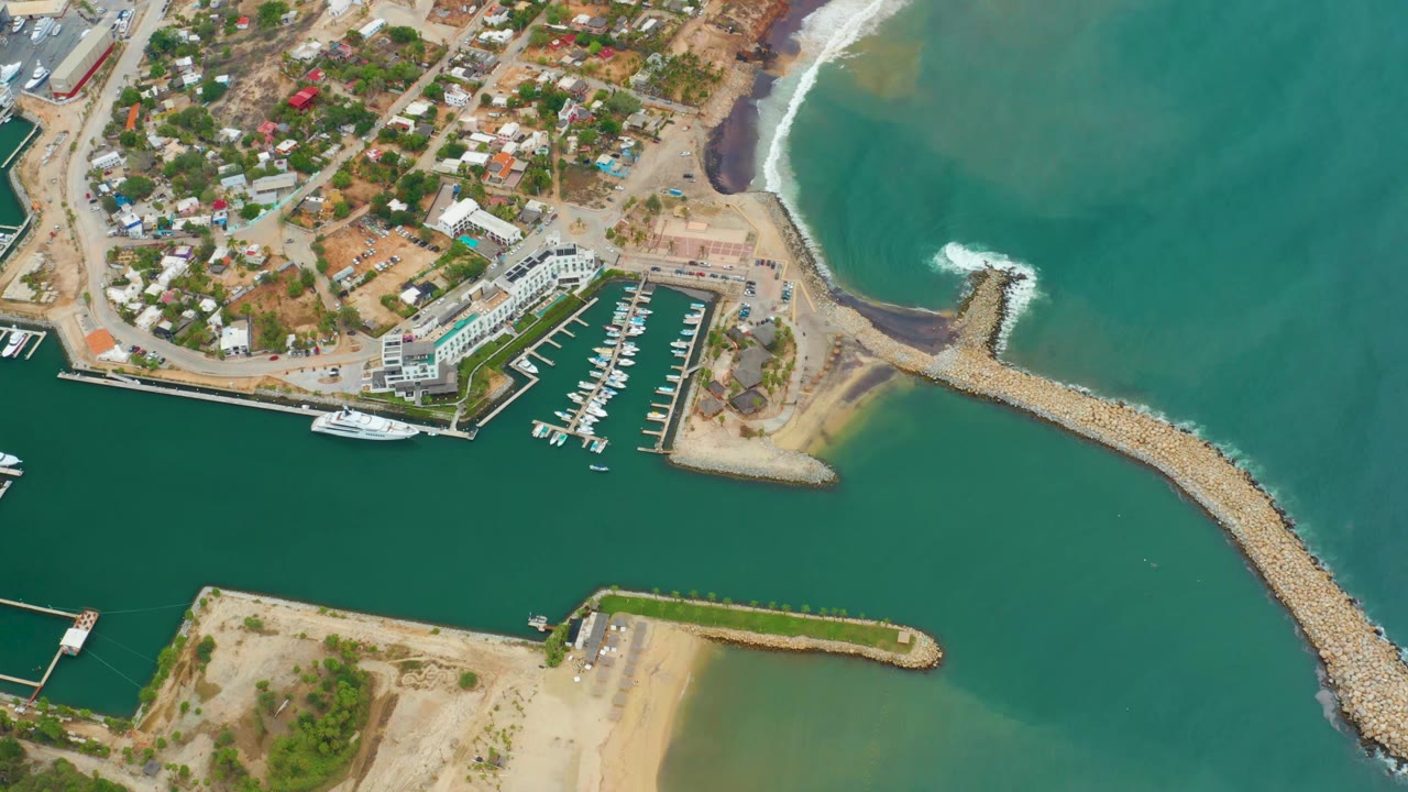 Aerial coastline frame with marina, homes, harbor water, jetty, and sandy foreground land.