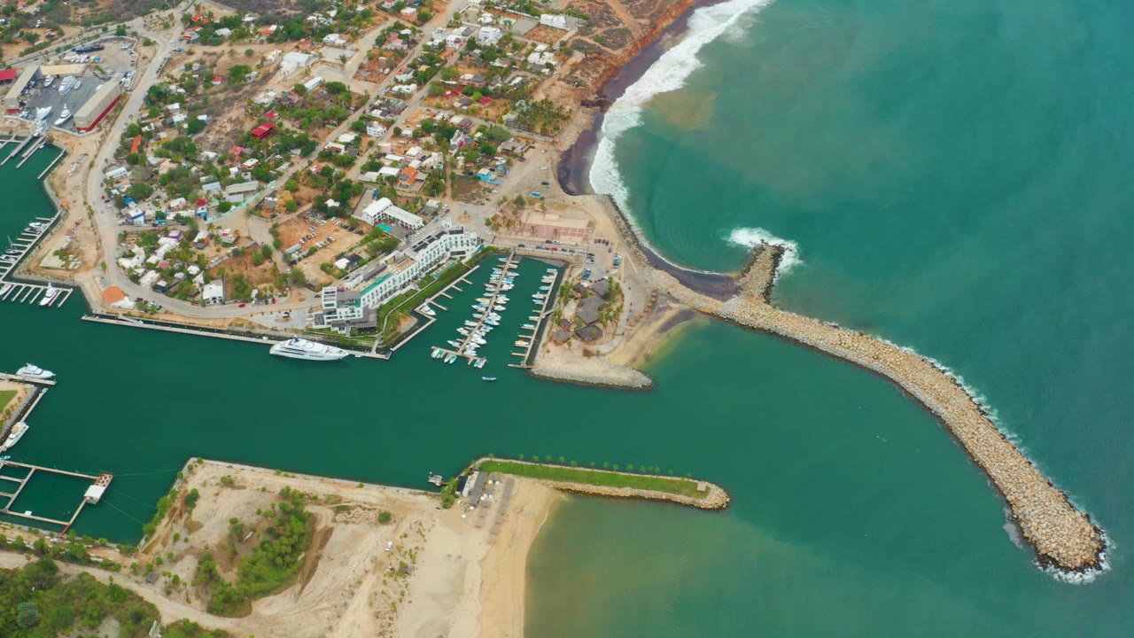 Aerial frame of La Playita Los Cabos showing the marina slips, curved rocky breakwater, sandy shoreline, turquoise water, coastal buildings, and no visible people.