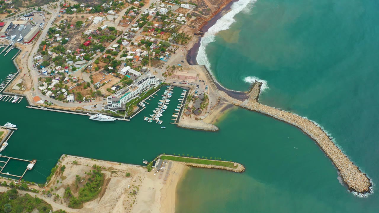 Drone-style aerial frame of La Playita Los Cabos showing green harbor water, docked boats, the diagonal rock breakwater, ocean swell near the shore, and no visible people.