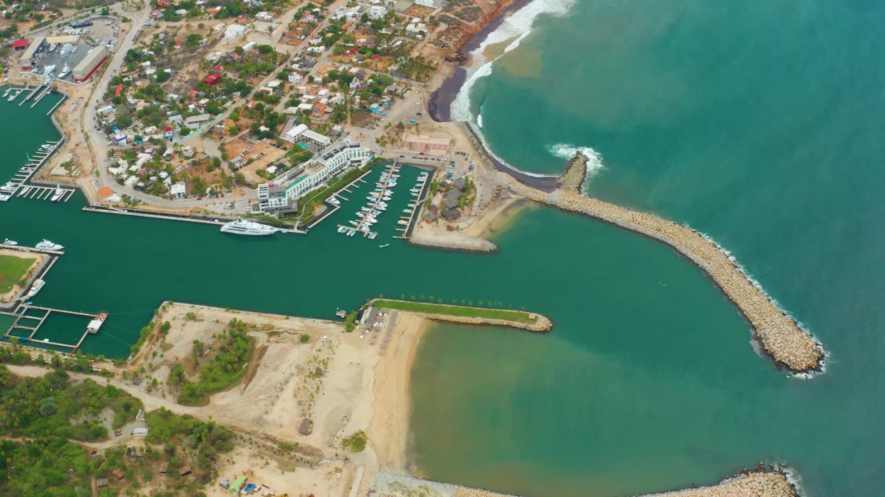 Opening aerial frame over La Playita Los Cabos showing the marina basin, sandy beach, rocky breakwater, coastal buildings, turquoise water, and no visible people.