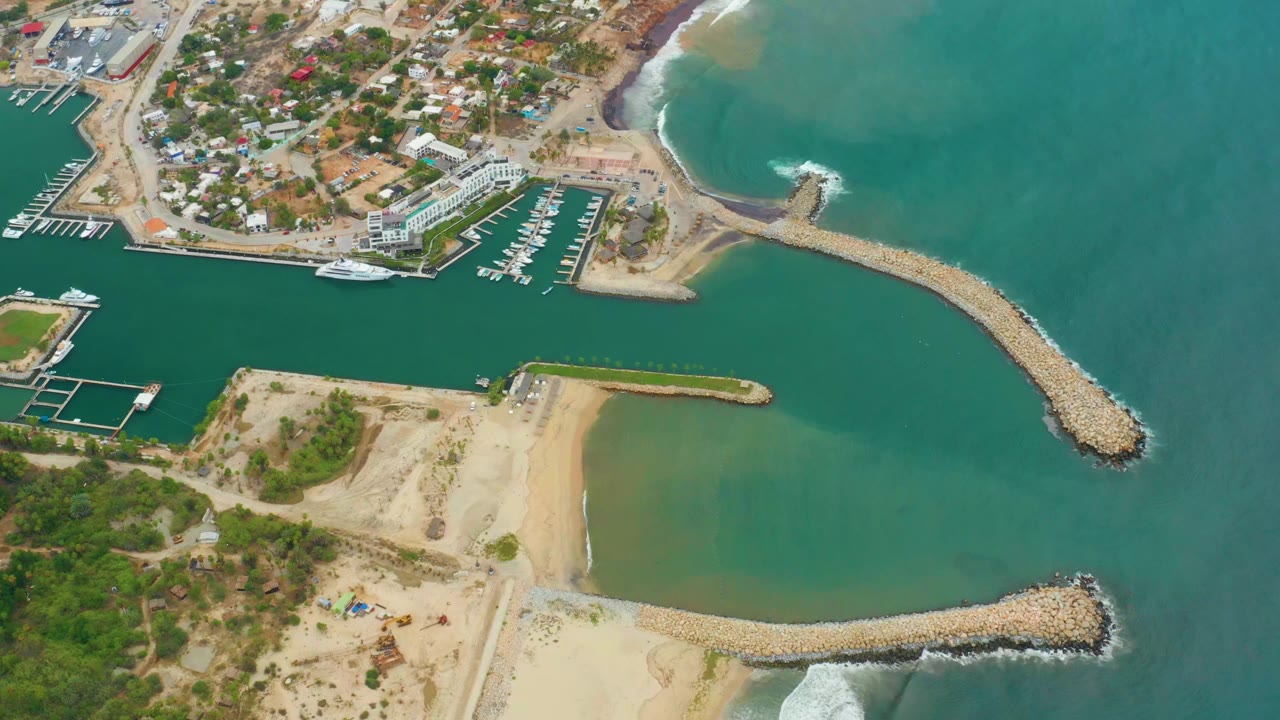 Aerial frame over La Playita Los Cabos showing marina docks at left, sandy beach below, curved rock breakwaters, turquoise harbor water, open ocean, and no visible people.