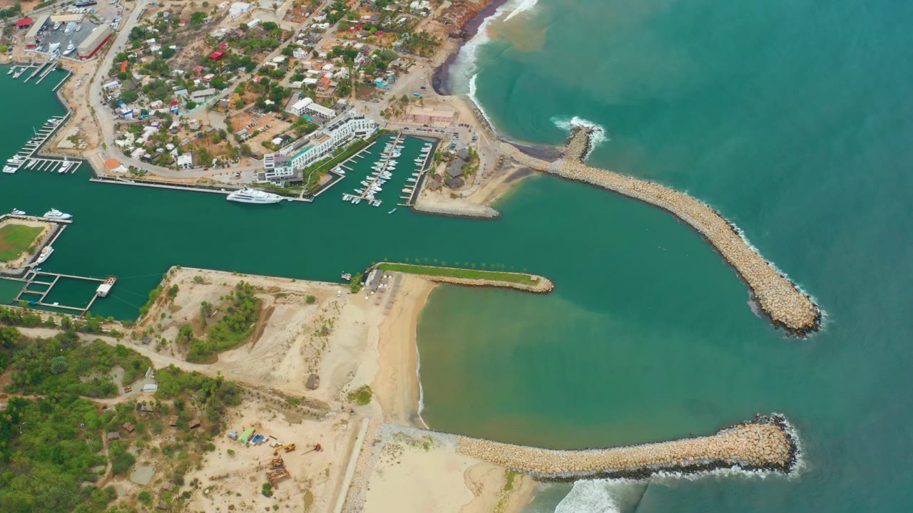 High aerial frame of La Playita Los Cabos showing dock rows, a white boat near the channel, pale beach sand, rock jetties, teal ocean water, and no visible people.