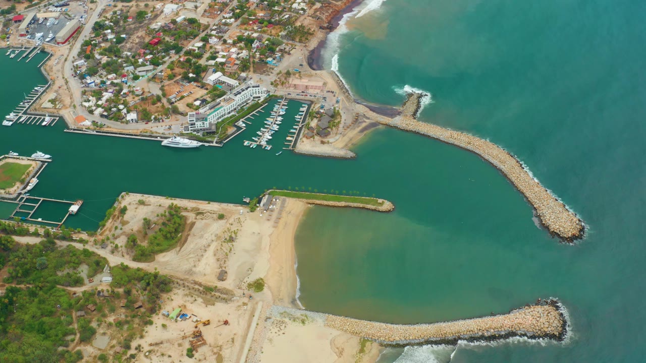 Aerial harbor entrance with marina, sandy shoreline, breakwater, and surf outside.