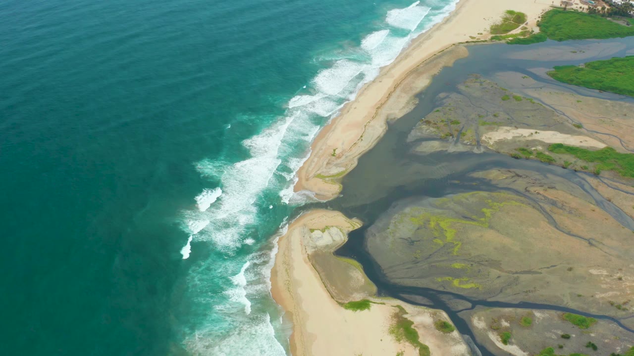 Aerial frame over La Playita Los Cabos places the breaking wave line near center, with dark river water and wet sand flats spreading to the right and no visible people.
