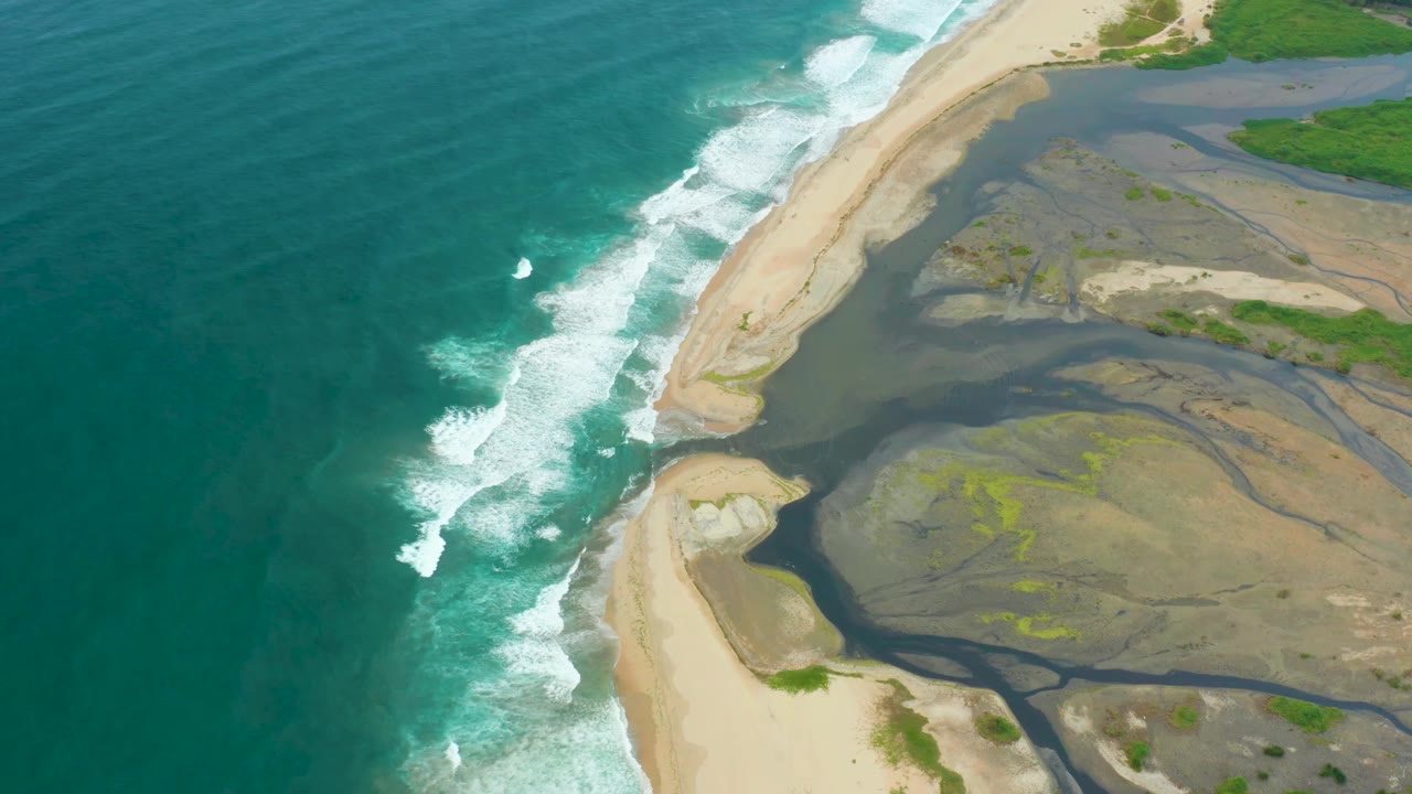 Aerial frame over La Playita Los Cabos keeps the ocean broad on the left, with foamy surf meeting the sandy mouth and darker estuary water behind it, no visible people.
