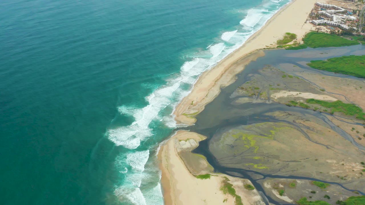 Aerial view of La Playita Los Cabos with turquoise ocean on the left, a sandy river mouth in the center, green vegetation at upper right, and no visible people.