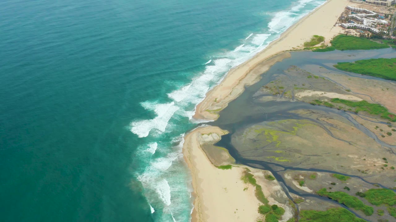 Aerial frame from La Playita Los Cabos showing turquoise ocean on the left, white surf at the beach, a dark estuary mouth across the sand, green patches inland, and no visible people.