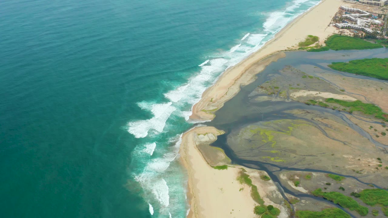High aerial frame from La Playita Los Cabos showing white wave foam curling toward the sandy shoreline, broad estuary flats to the right, distant coastal buildings at the top, and no visible people.
