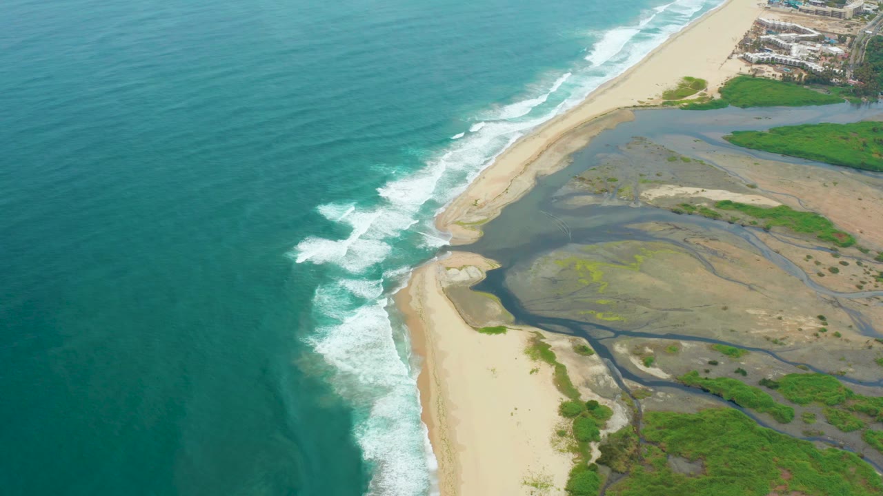 Aerial view over La Playita Los Cabos with turquoise ocean on the left, white surf on the sand, a dark estuary channel, green vegetation, and no visible people.
