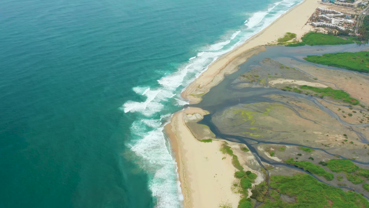 Aerial view in La Playita Los Cabos with the ocean occupying the left half, white waves reaching the beach, braided estuary water on the right, and no visible people.