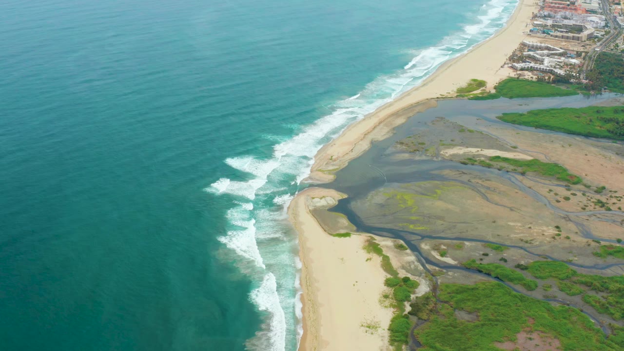 Aerial view over La Playita Los Cabos with teal ocean at left, white surf on pale sand, wetland channels inland, distant buildings at the top edge, and no visible people.