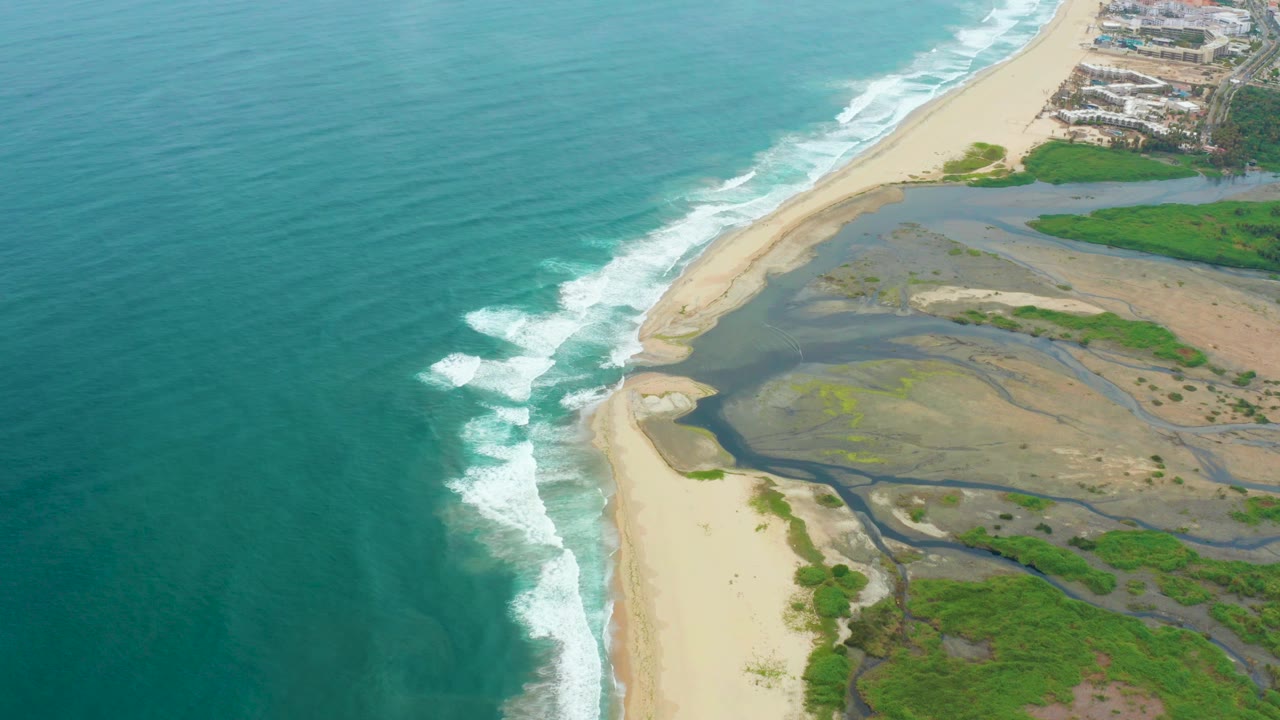 Surf breaks along a pale beach next to shallow estuary water.