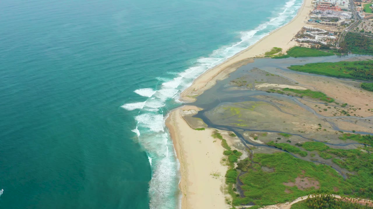 Aerial frame from La Playita Los Cabos, Mexico with the surf line slightly farther along the sand, the estuary mouth visible near the beach, green vegetation below, upper-right buildings, and no visible people.