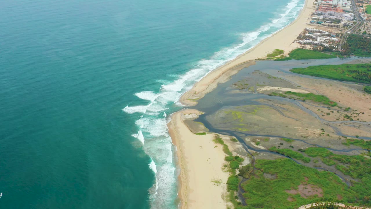 High aerial view of waves, beach, estuary channels, dunes, vegetation, and coastal buildings.