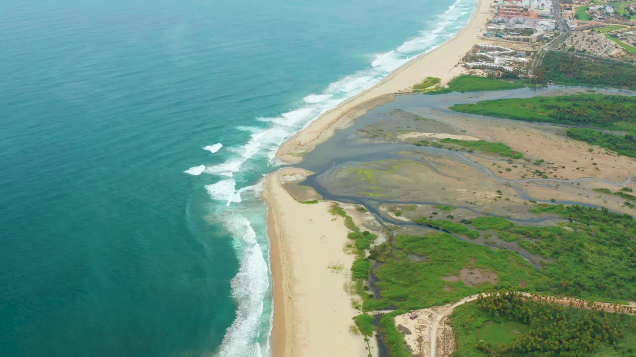 Aerial view of La Playita Los Cabos with teal ocean on the left, white surf curling toward the beach, lagoon flats beside green vegetation, distant buildings, and no visible people.
