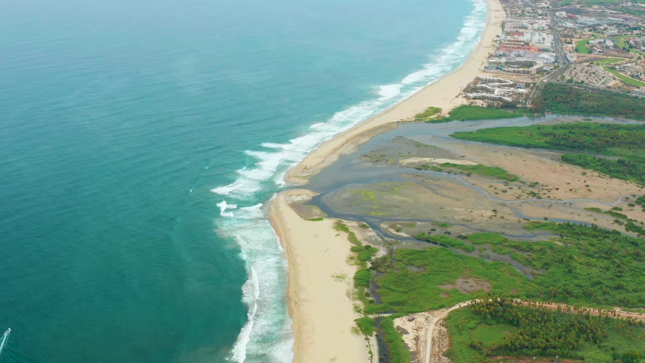 Aerial view over La Playita Los Cabos showing turquoise ocean, white surf on a sandy beach, inland wetlands, green vegetation, a small coastal settlement near the top edge, and no visible people.
