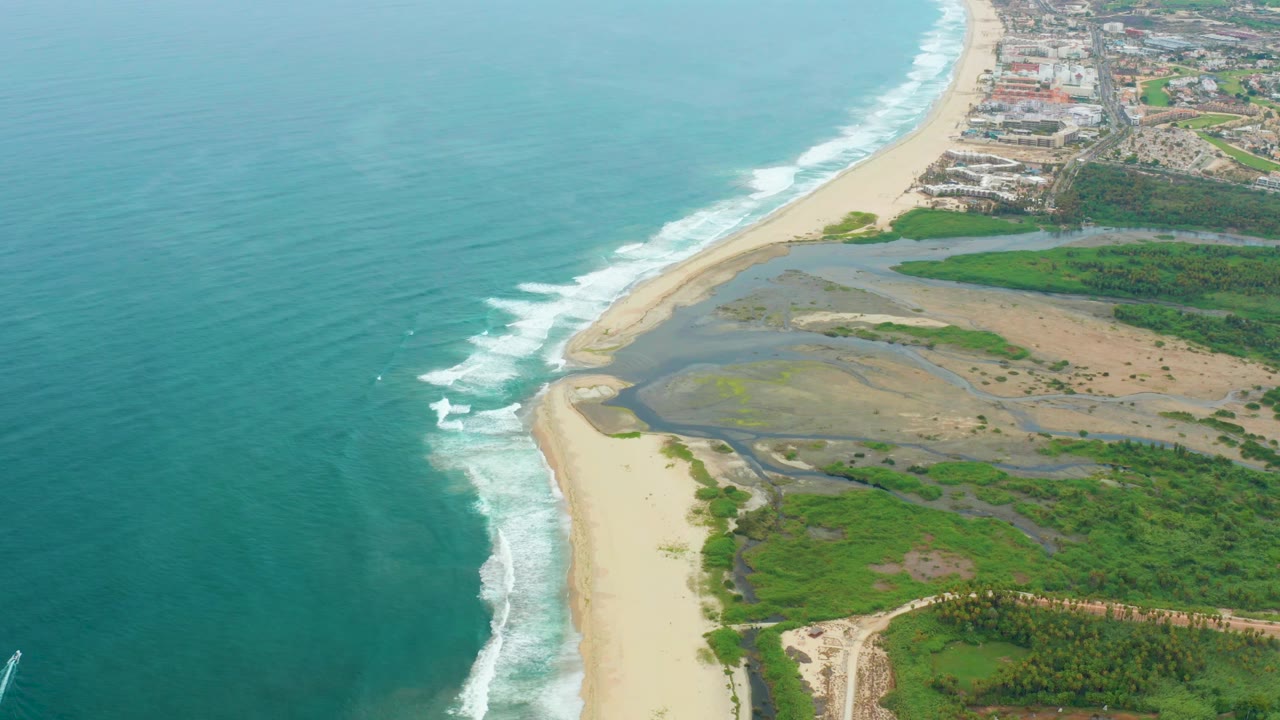 Aerial frame from La Playita Los Cabos with broad turquoise water, a curving foam line on the beach, wetland channels inland, dense green vegetation, distant buildings, and no visible people.