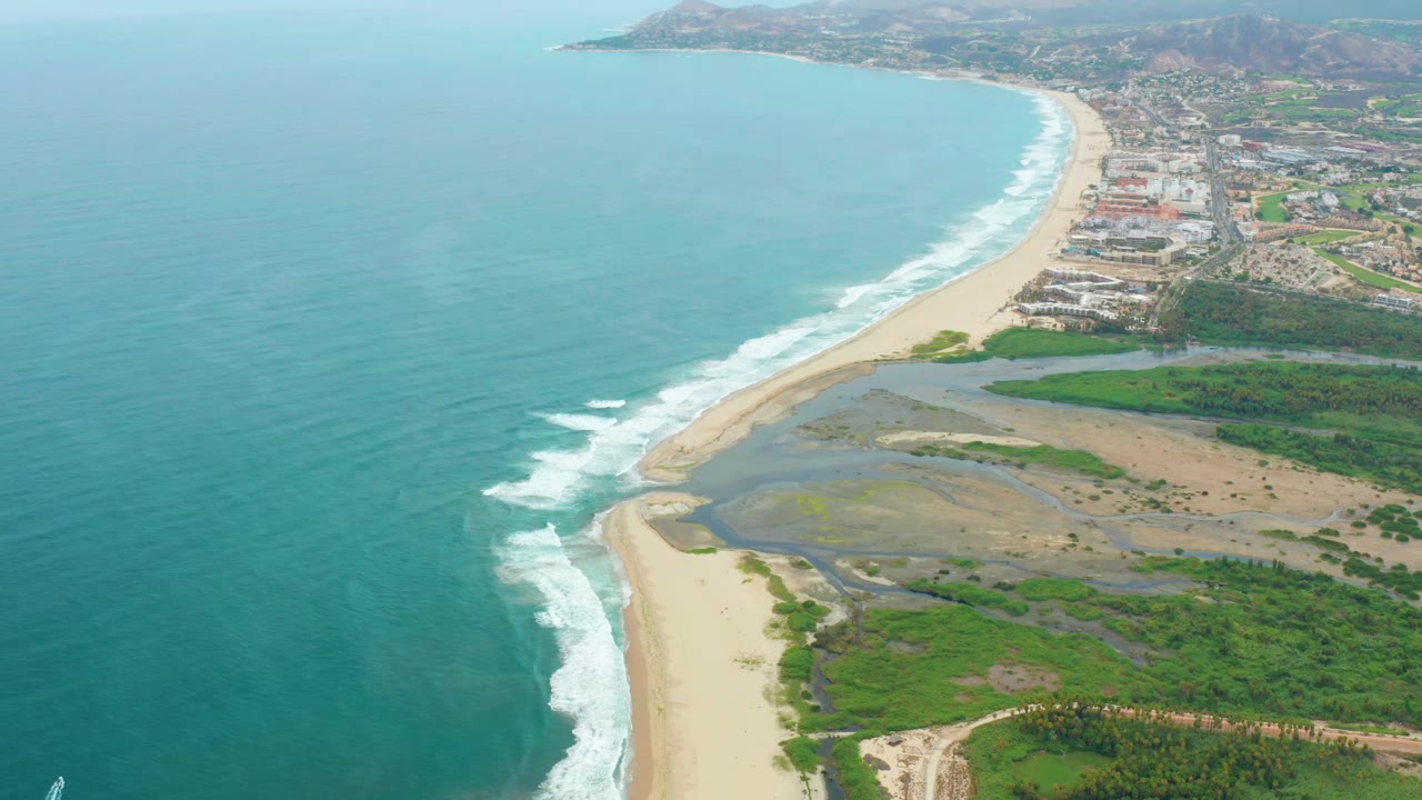 Aerial frame over La Playita Los Cabos showing surf bands advancing along the curved beach, lagoon shallows to the right, dense green patches inland, distant coastal buildings, and no visible people.