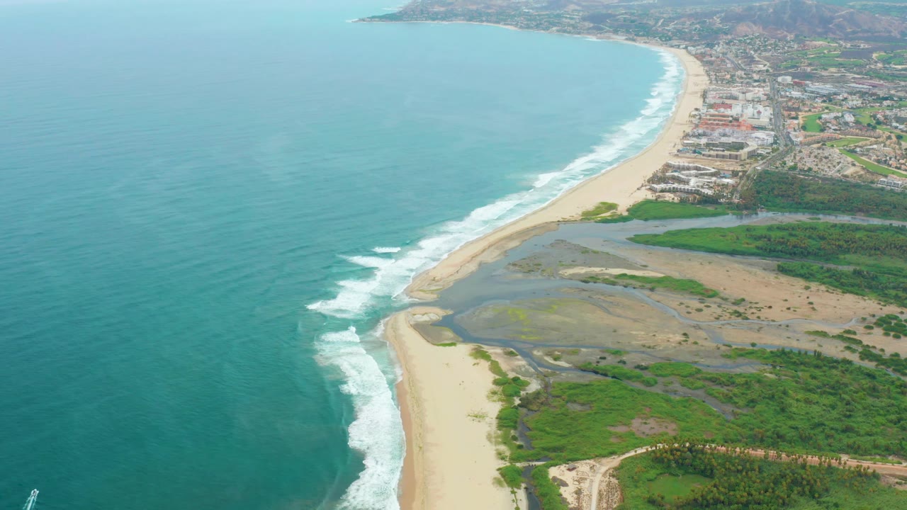 High drone view in La Playita Los Cabos with more open ocean visible to the left, waves breaking in white strips, sandy beach and lagoon through the middle, coastal town beyond, and no visible people.