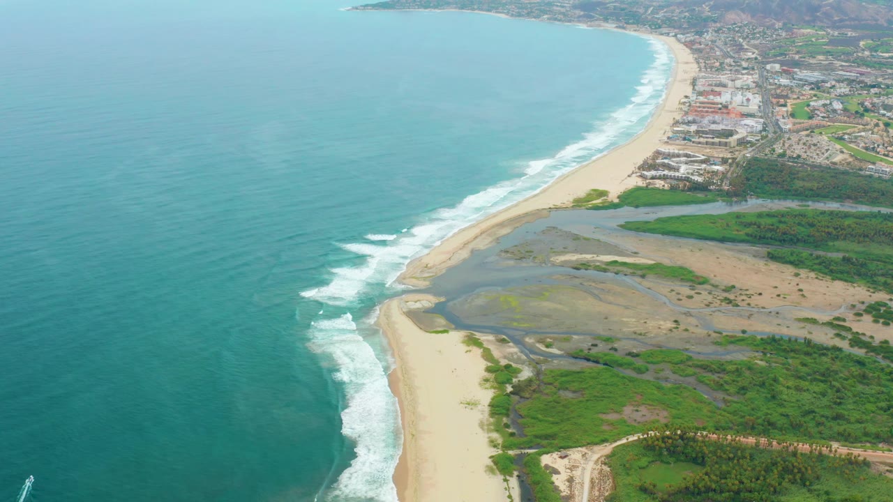 Mid-clip aerial over La Playita Los Cabos showing broad teal water, surf hitting a sandy spit, lagoon pools inland, green coastal vegetation, background buildings, and no visible people.