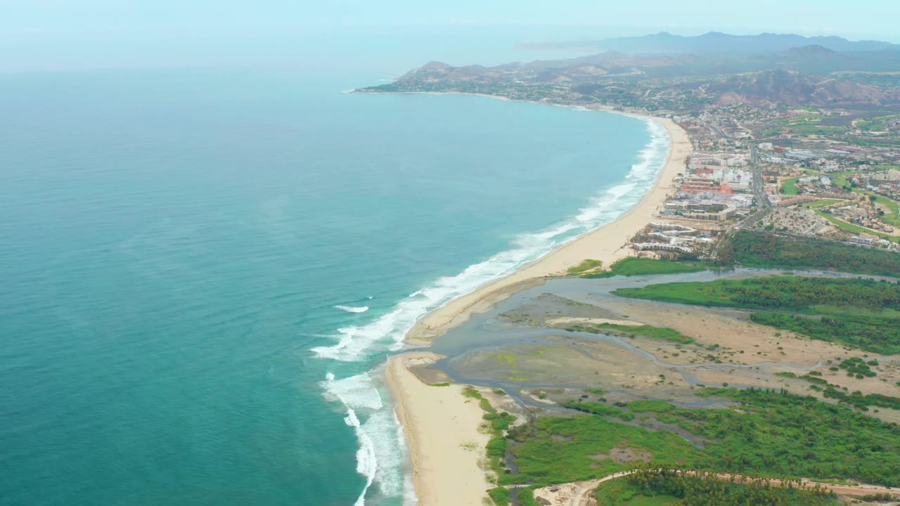 Aerial view over La Playita Los Cabos showing a broad blue ocean, breaking waves curling toward the sand, low buildings behind the beach, green patches near shore, and no visible people.