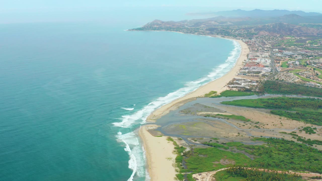 Aerial frame from La Playita Los Cabos with the coastline running diagonally, turquoise water filling the left side, foamy waves along the beach, town buildings inland, and no visible people.