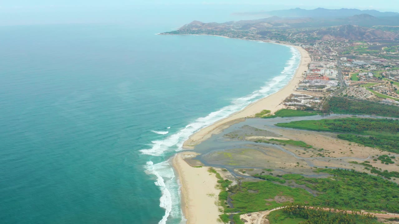 Coastal aerial showing open ocean, surf, sand flats, greenery, and buildings.
